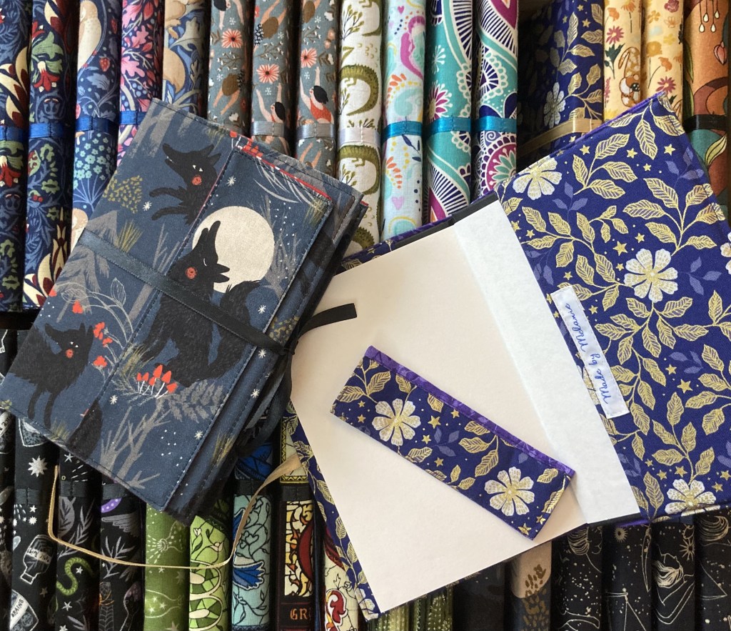 Image showing a closed notebook with a wolf on the cover and an opened notebook with white flowers and a matching bookmark laid on top of many fabric covered notebooks spines up